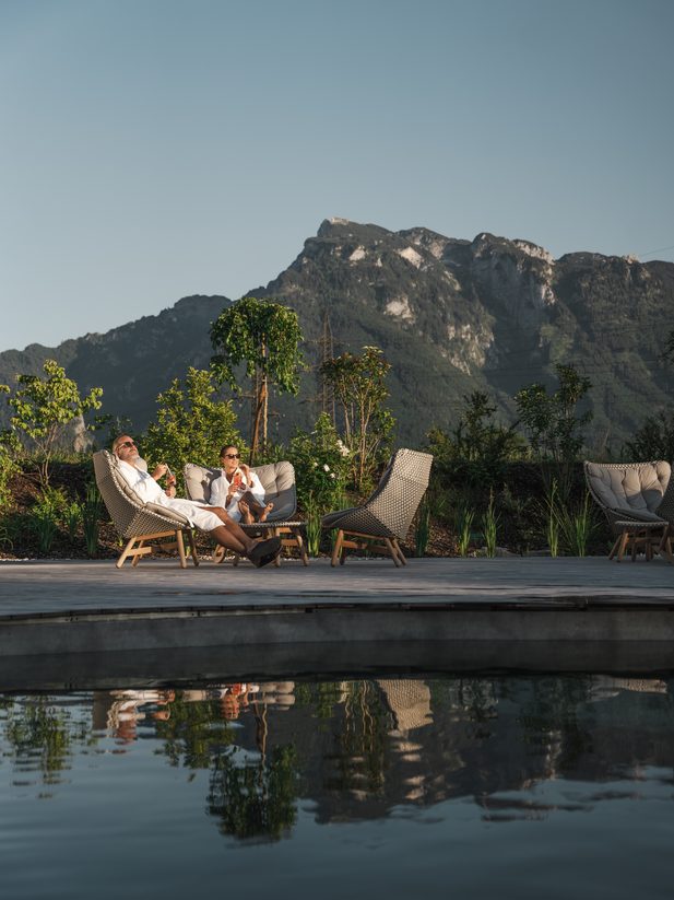 Zwei Gäste im Bademantel entspannen auf Liegestühlen am Pool mit Bergpanorama im Hintergrund.