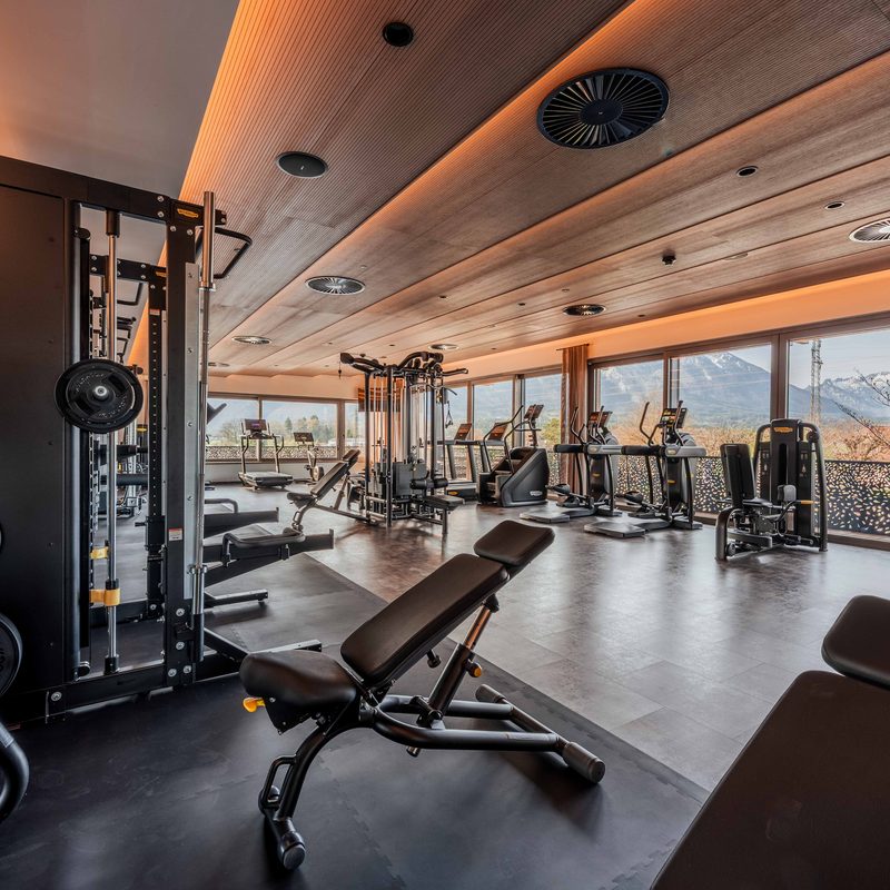 Fitnessraum mit Kraft- und Cardiogeräten vor großen Fenstern mit Blick auf die Berge.