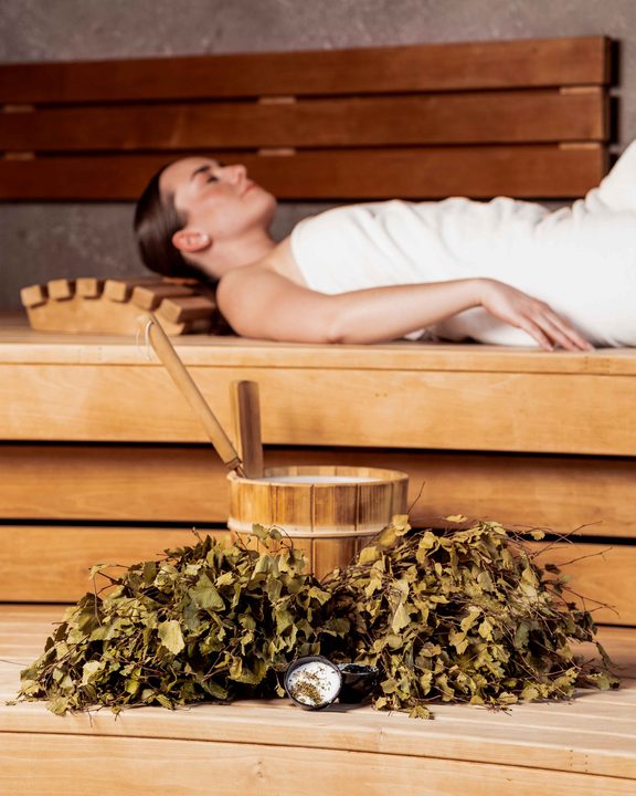 Woman lying on a sauna bench with an infusion bucket, ladle and birch branches in front.