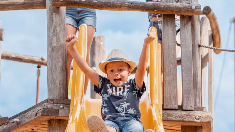 Junge rutscht eine gelbe Rutsche hinunter, zwei Mädchen jubeln auf dem Holzspielplatz.