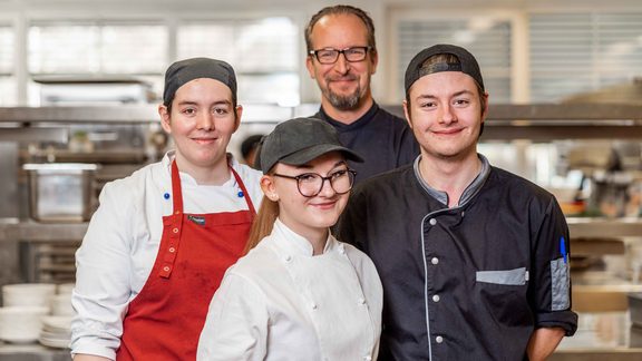 Four kitchen staff stand in a restaurant kitchen and look at the camera.