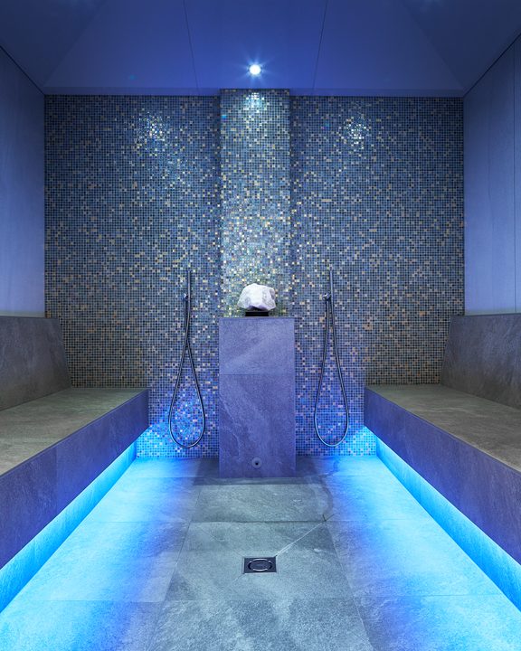 Modern hammam steam room with mosaic wall, stone benches and blue LED lighting.
