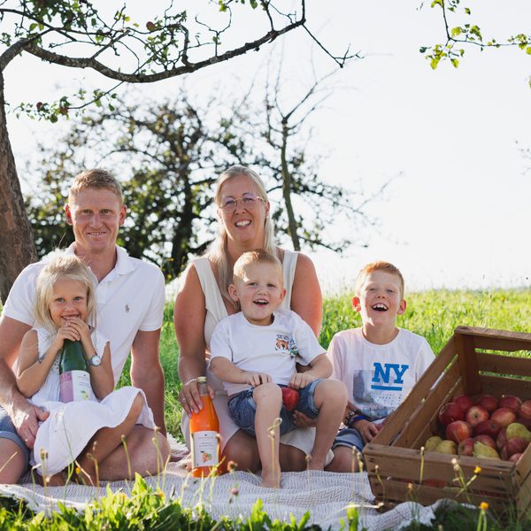Familie auf Picknickdecke im Obstgarten neben Apfelkiste und Saftflaschen.
