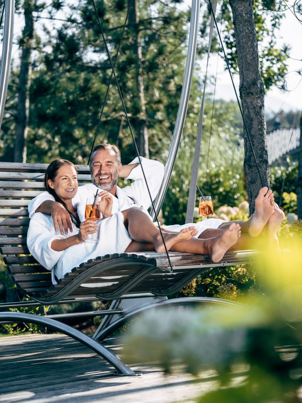 Paar im Bademantel auf Schwingliege im Garten, mit zwei Drinks in der Hand.