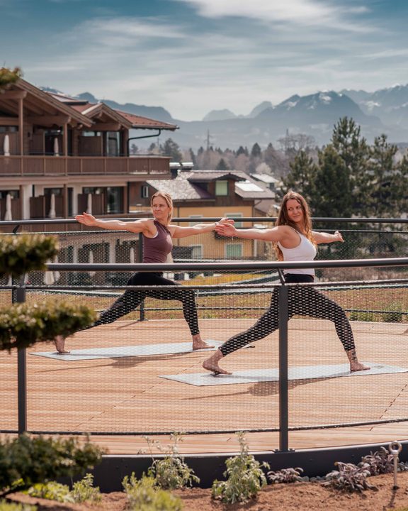 Zwei Frauen machen Yoga auf einer Outdoor-Terrasse mit Bergblick.