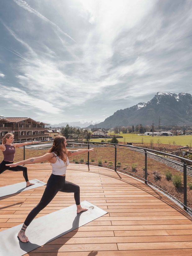 Zwei Frauen machen Yoga auf einer Hotelterrasse mit Blick auf Berge und weite Landschaft.