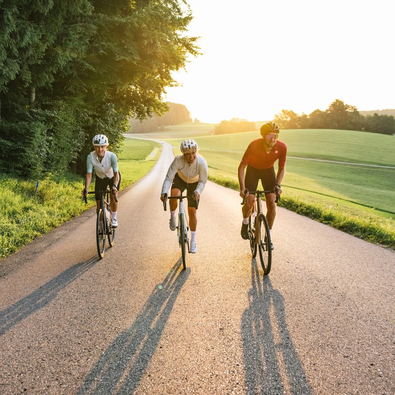 Three cyclists on road bikes ride along a country road through rolling fields in evening light.