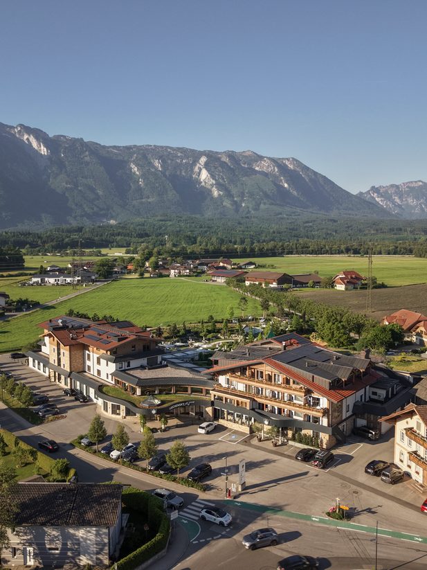 Luftaufnahme einer Hotelanlage mit Parkplatz vor grünen Wiesen und einem Alpenmassiv im Hintergrund.