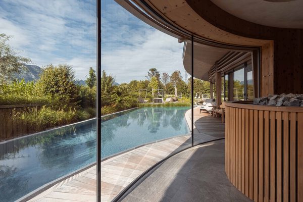 Curved indoor pool with glass wall and loungers, looking out to the garden and mountains.