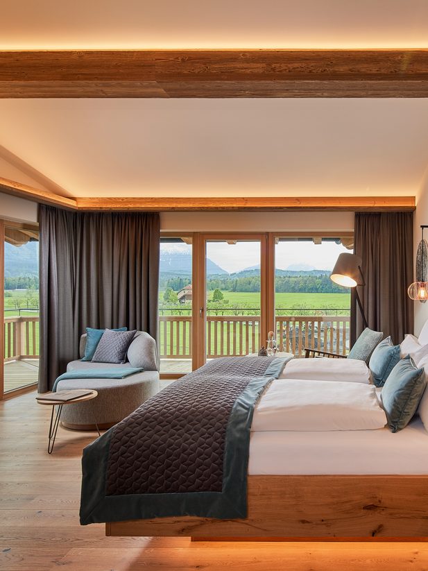 Modernes Hotelzimmer mit Doppelbett, Sitzecke und Fensterfront zur Terrasse mit Blick auf Wiesen und Berge.